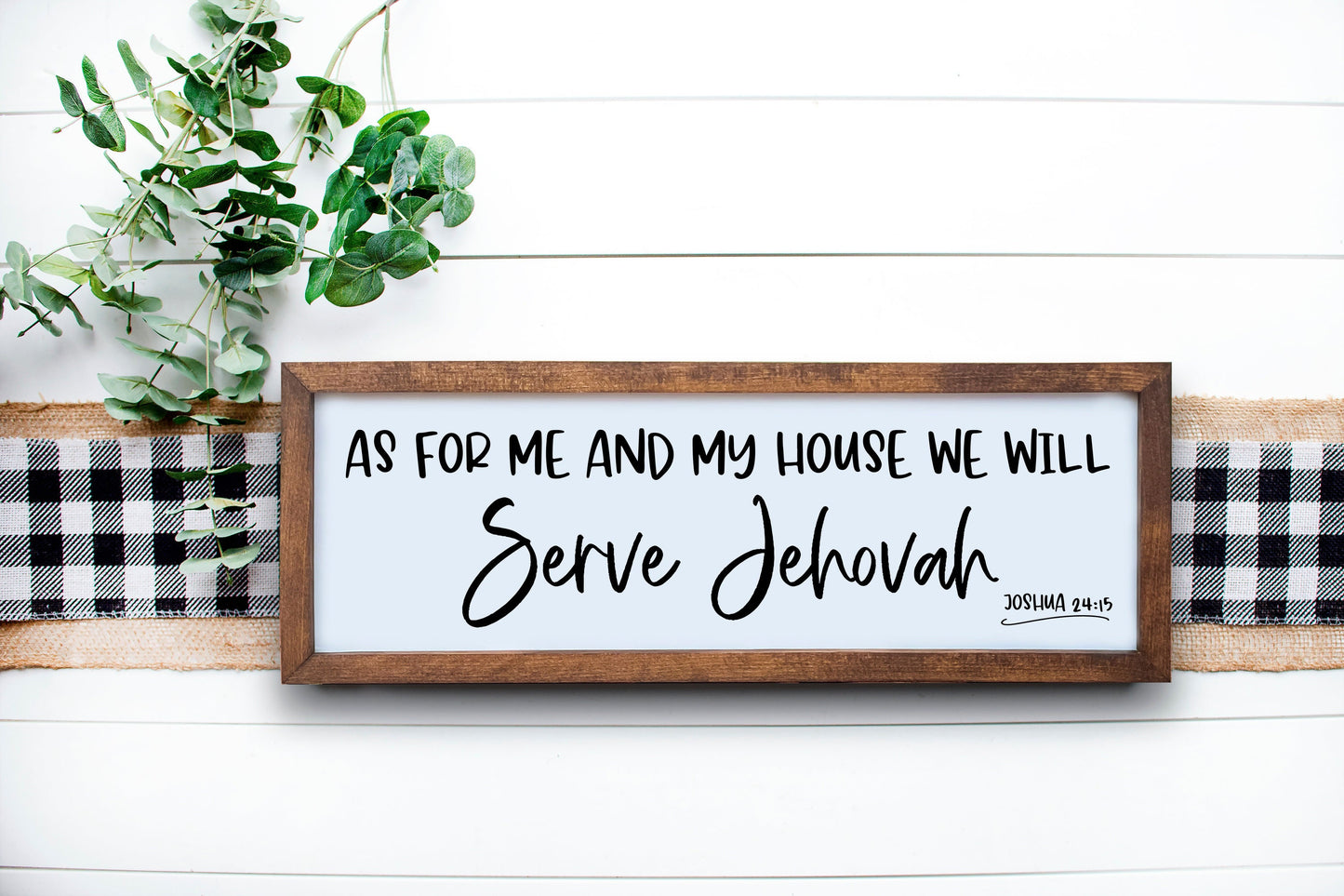 A wooden sign with a Bible verse 'As for me and my house, we will serve Jehovah' written on it, placed on a white wooden surface with a green plant and a checkered pattern in the background.